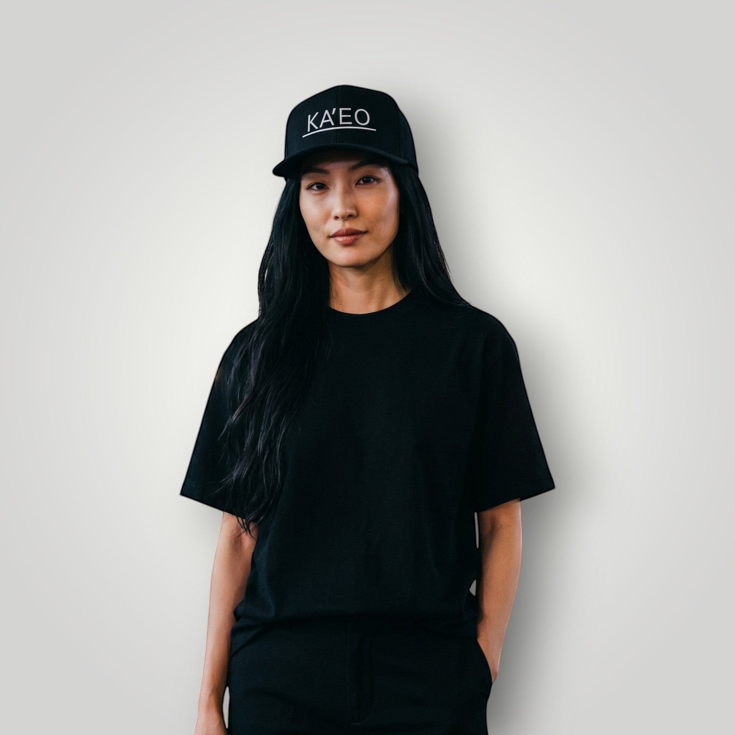 KA'EO Apparel black embroidered snapback cap worn by a woman in a city at night.