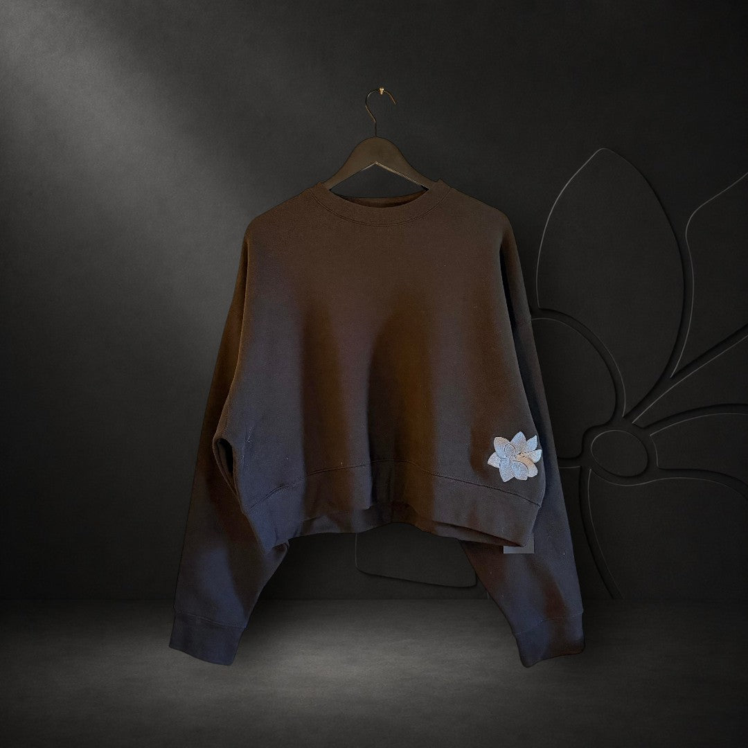 KA'EO Apparel black relaxed cropped fleece crewneck sweatshirt with embroidered Sampa flower detail on the hem.