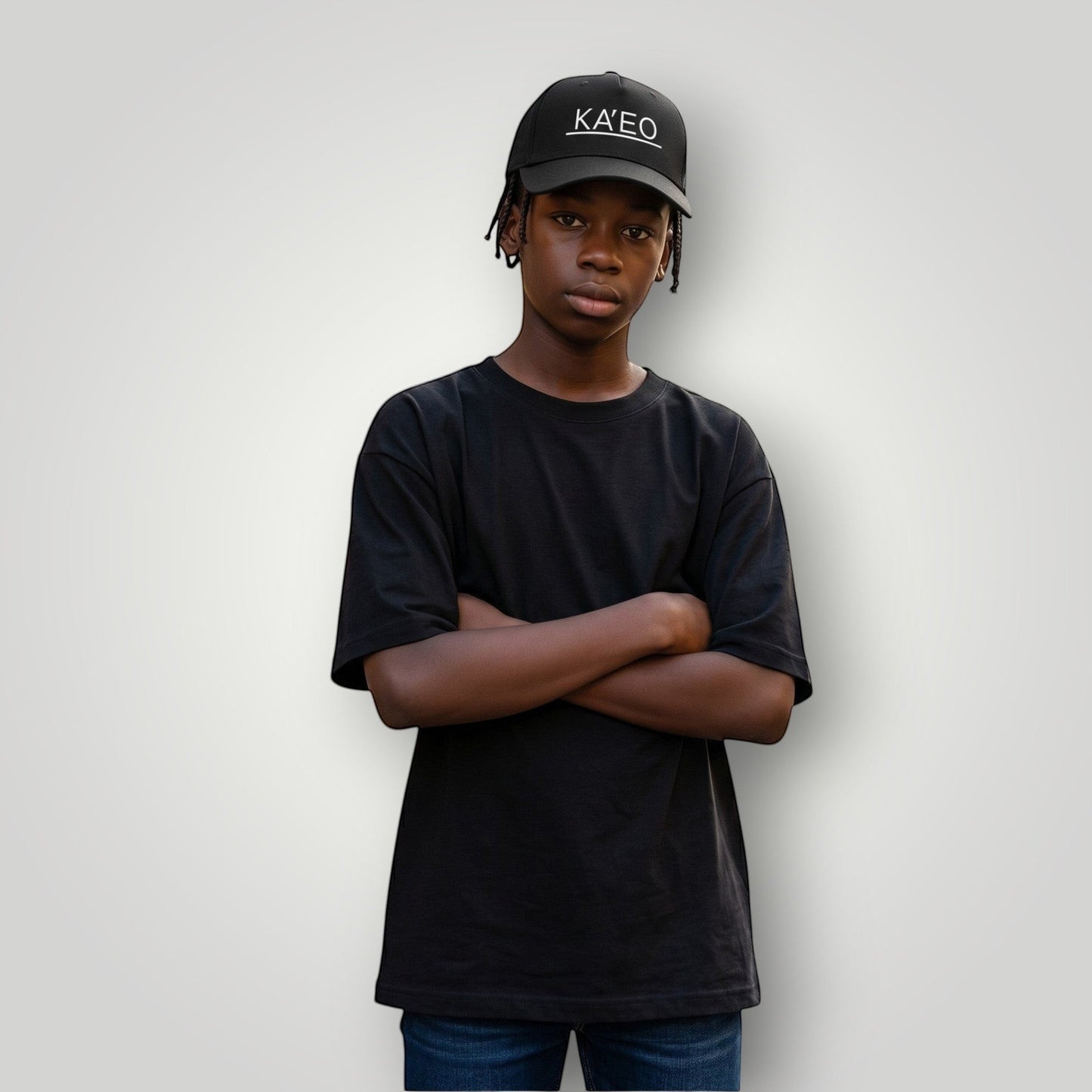 Black KA'EO Apparel embroidered snapback cap worn by a young Black male with arms crossed, standing on a street.