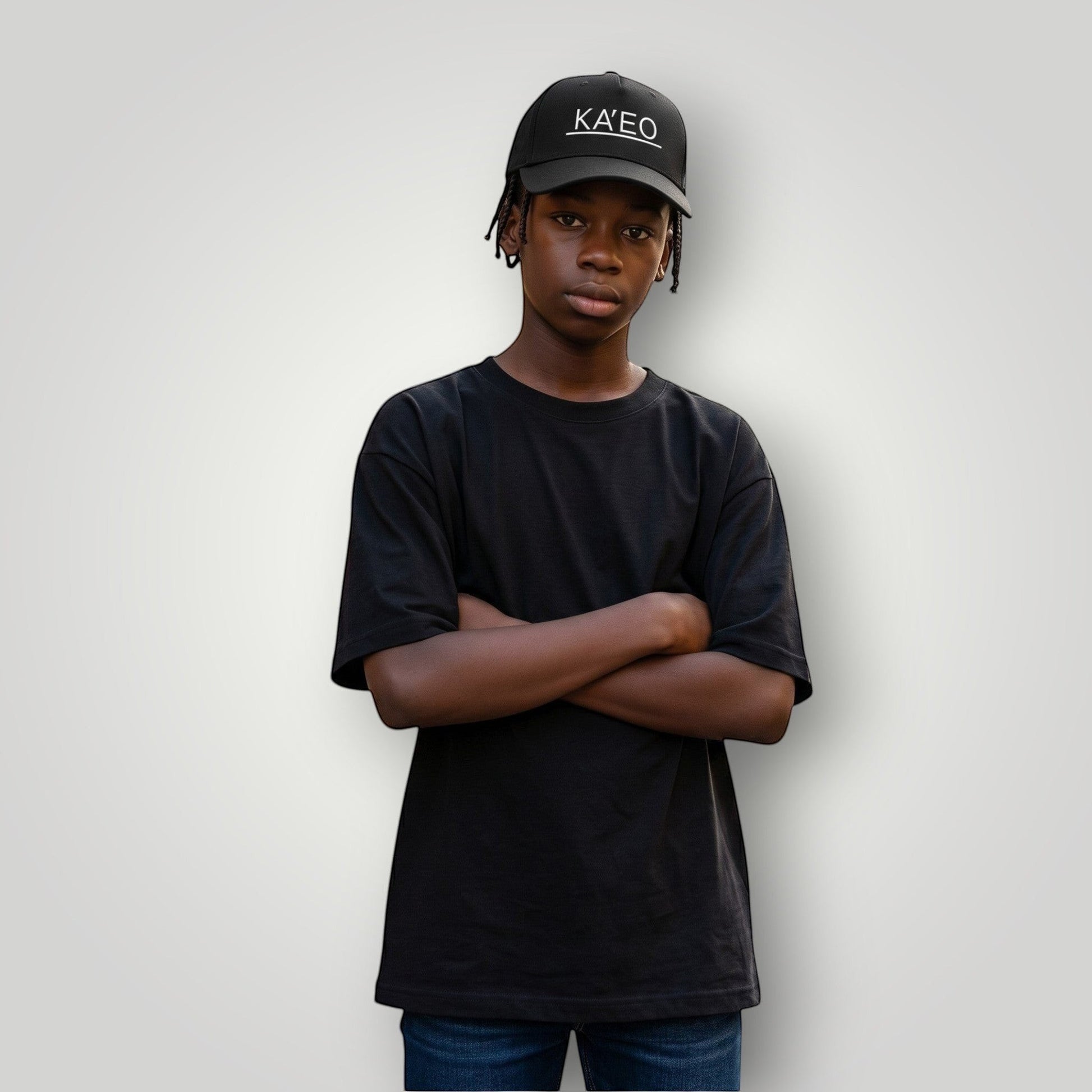 Black KA'EO Apparel embroidered snapback cap worn by a young Black male with arms crossed, standing on a street.