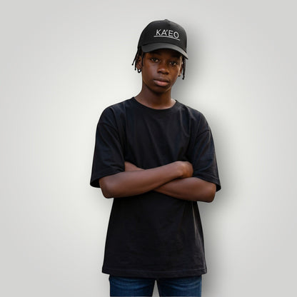 Black KA'EO Apparel embroidered snapback cap worn by a young Black male with arms crossed, standing on a street.