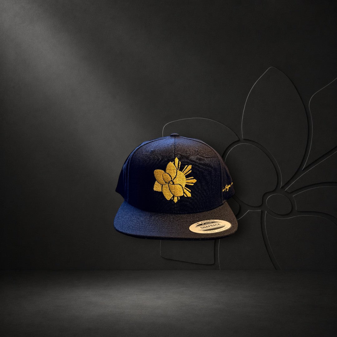 KA'EO Apparel black snapback cap featuring a gold embroidered sampaguita flower design and flat brim.