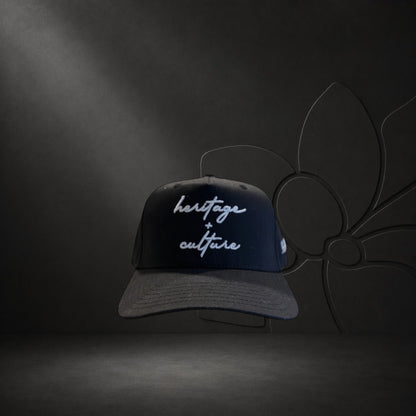 KA'EO Apparel black cotton snapback cap with embroidered 'heritage + culture' text and curved brim.